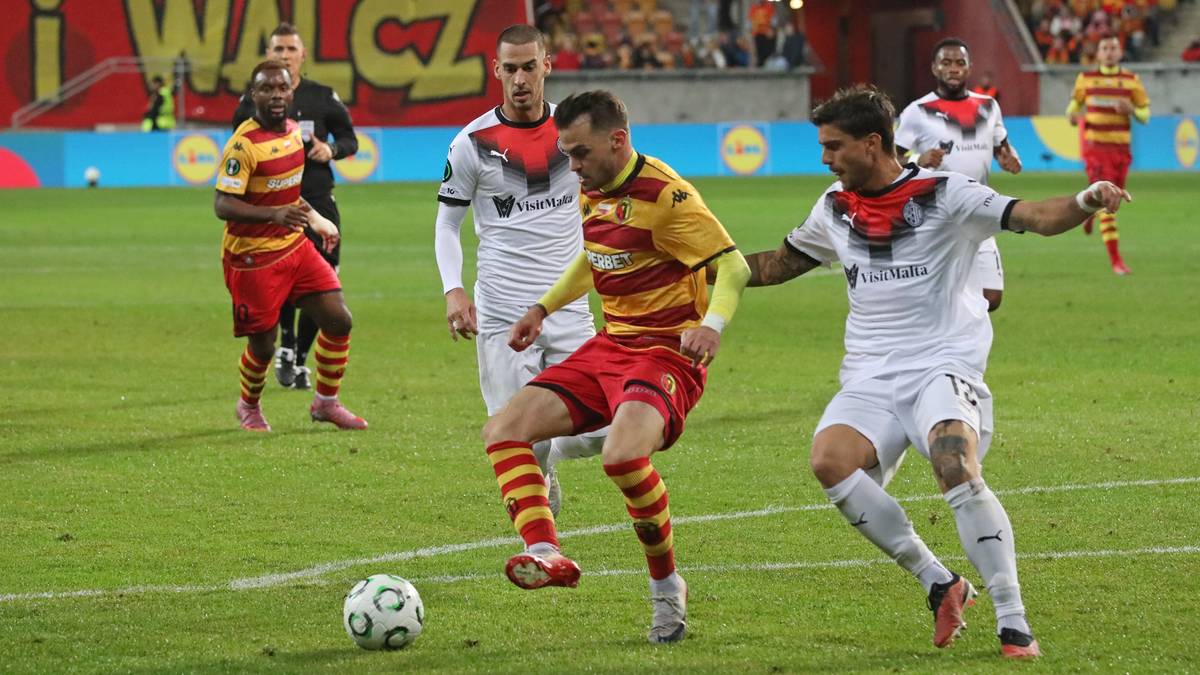 Piłkarze w czerwono-żółtych i białych strojach rywalizują o piłkę na boisku piłkarskim.