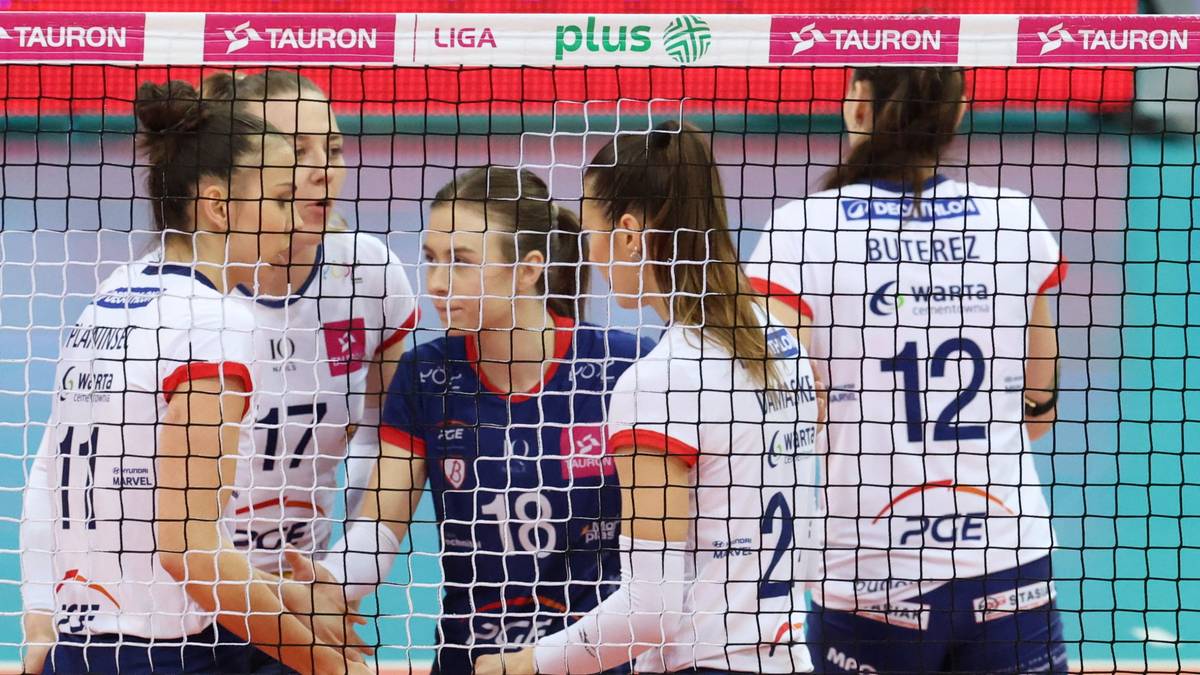 Cztery siatkarki w białych strojach sportowych stoją blisko siebie, otoczone siatką, z widocznymi numerami na koszulkach.