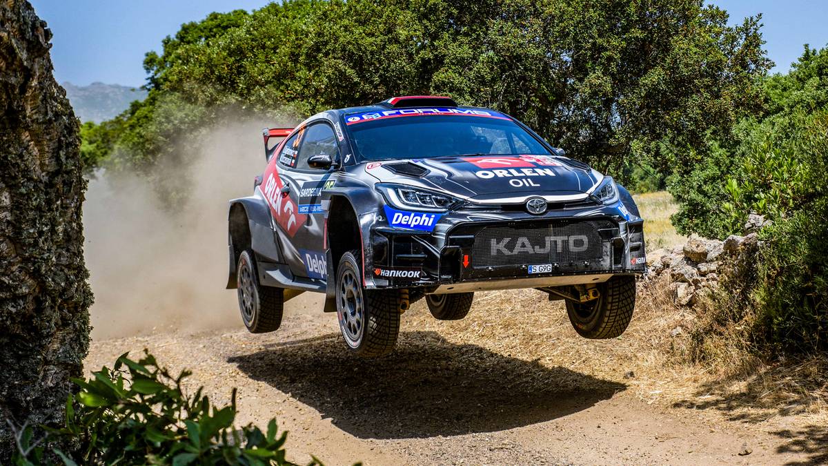 Samochód rajdowy z napisem KAJTO, w barwach ORLEN OIL, lekko unoszący się nad ziemią na zakurzonym, szutrowym zakręcie, w otoczeniu zielonych drzew i krzewów.