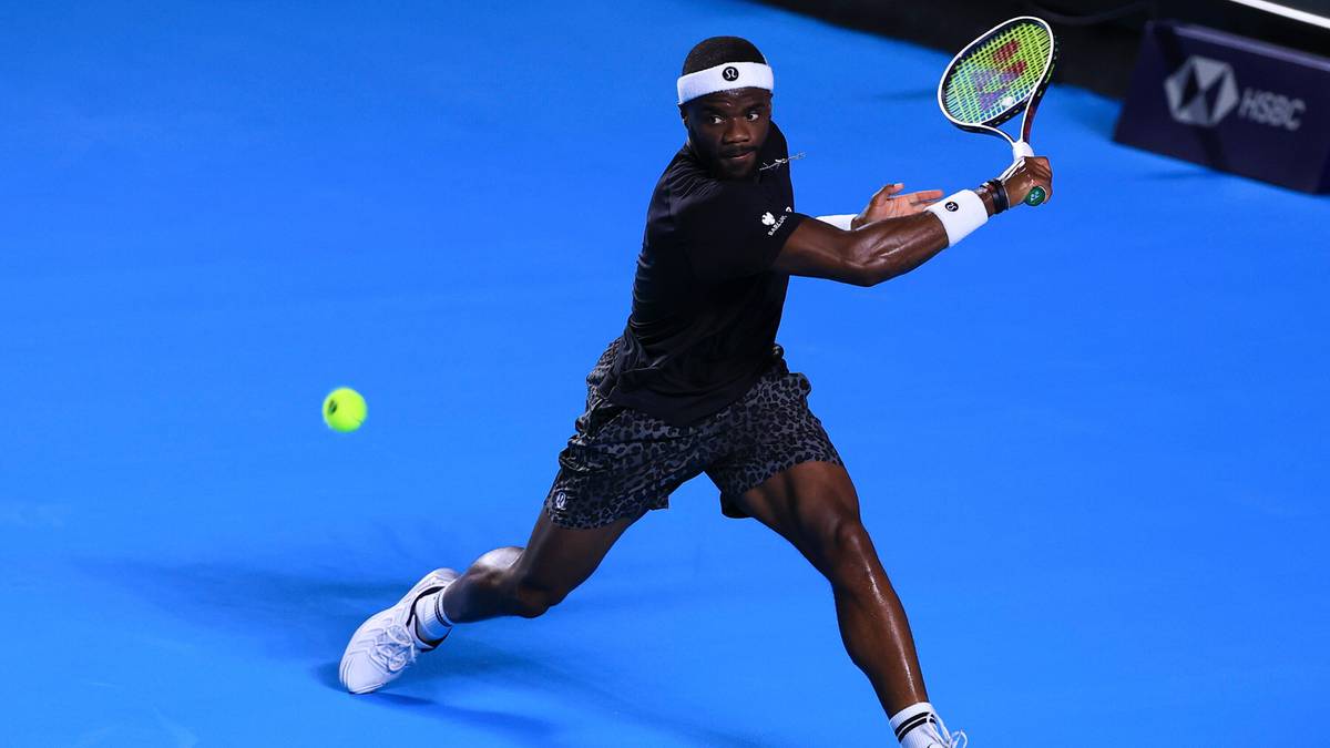 Tenisista Frances Tiafoe w czarnym stroju sportowym uderza piłkę forhendem na niebieskim korcie tenisowym.