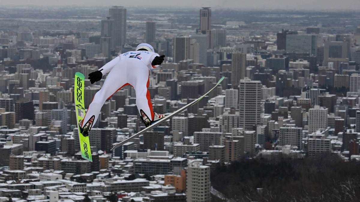 Skoczek narciarski w białym stroju z zielonymi nartami w locie nad zabudowanym miastem.