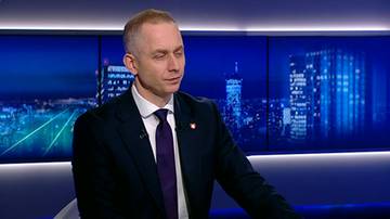 Mężczyzna w ciemnym garniturze i fioletowym krawacie siedzi na krześle. Za nim znajduje się niebieski ekran z podświetlonymi sylwetkami budynk&oacute;w.