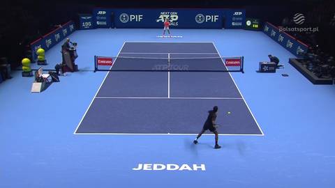 Dw&oacute;ch tenisist&oacute;w grających mecz na niebieskim korcie tenisowym. Jeden z nich, w ciemniejszym stroju, odbija piłkę w kierunku przeciwnika, kt&oacute;ry stoi bliżej siatki.