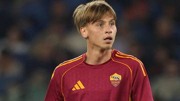 Młody mężczyzna w koszulce piłkarskiej AS Roma z herbem klubu na piersi i logo Adidasa, z blond włosami, patrzy w bok z otwartymi ustami.