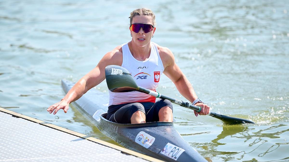 Zawodnik kajakarstwa w akcji, ubrany w białą koszulkę z polskim godłem, wiosłuje na jeziorze podczas zawodów.