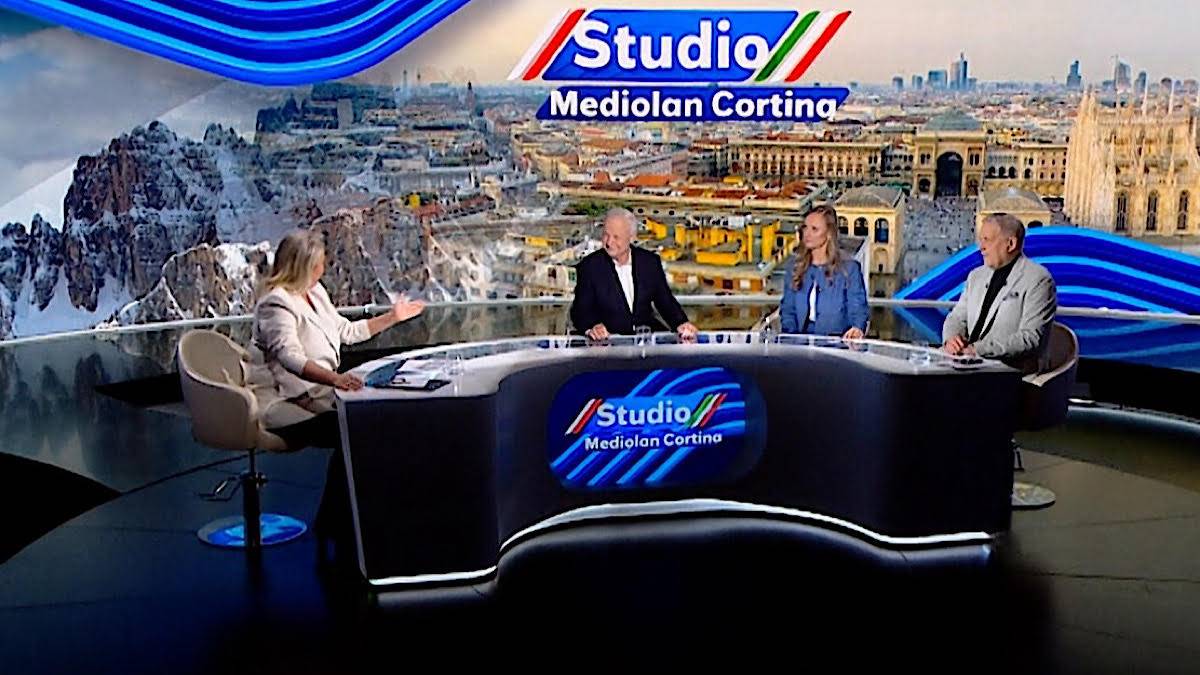 Cztery osoby siedzą w studiu telewizyjnym przy stole. W tle znajduje się grafika przedstawiająca panoramę miasta z g&oacute;rami.