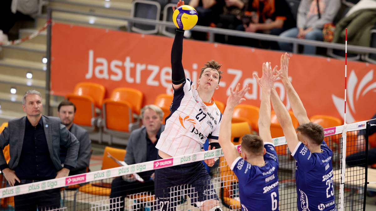 Siatkarz z numerem 27 na koszulce wykonuje zagrywkę, zapatrzony w piłkę unoszoną nad głową. Po drugiej stronie siatki widoczni są dwaj zawodnicy w niebieskich strojach, blokujący próbę zagrywki.