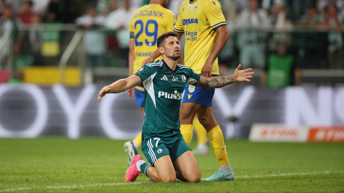 Piłkarz Legii Warszawa w zielonym stroju klęczy na boisku z wyciągniętymi rękami i wyrazem rozpaczy na twarzy.