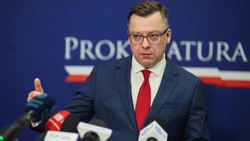 Mężczyzna w garniturze i czerwonym krawacie przemawia przy mównicy, za nim logo prokuratury.