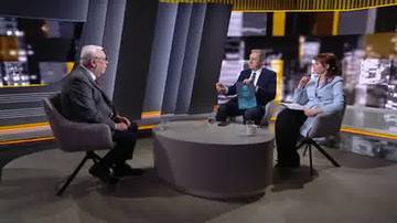 Trzy osoby siedzą przy okrągłym stoliku w studiu telewizyjnym. W środku siedzi mężczyzna w garniturze, po jego lewej stronie siedzi starszy mężczyzna w ciemnym garniturze, a po prawej stronie siedzi kobieta w jasnej marynarce.