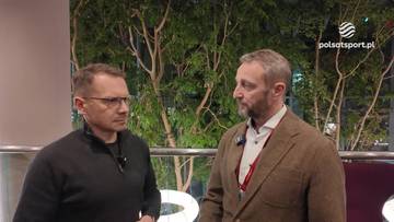 Dwóch mężczyzn rozmawia ze sobą, jeden ubrany w czarną bluzę, drugi w brązową marynarkę i białą koszulę. W tle widać drzewa.