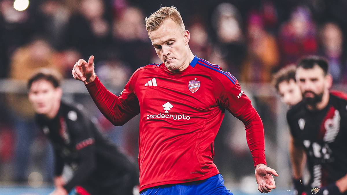 Piłkarz w czerwonej koszulce i niebieskich spodenkach kopie piłkę na zielonym boisku.