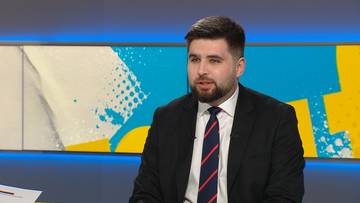 Mężczyzna z brodą w czarnym garniturze i krawacie w biało-granatowe pasy siedzi przed kolorowym tłem.