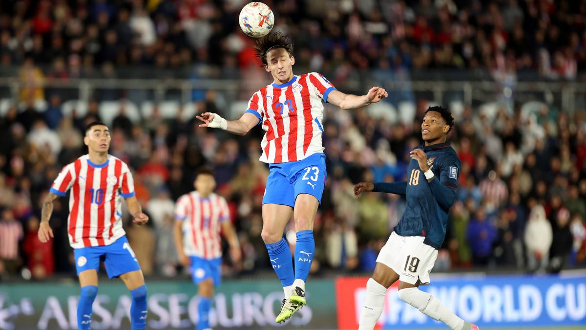 Piłkarz w biało-czerwonej koszulce wyskakuje do strzału głową, z piłką nad głową. Na pierwszym planie inny piłkarz w niebieskiej koszulce, biegnący do przodu. W tle widzowie na trybunach.