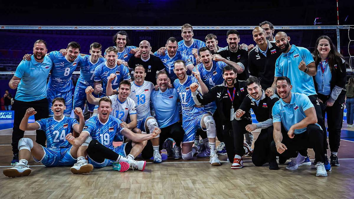 Uśmiechnięta drużyna siatkarzy w niebieskich strojach pozuje do zdjęcia po wygranym meczu.