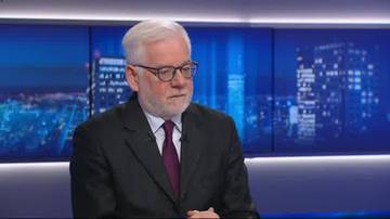 Starszy mężczyzna w okularach i garniturze, siedzący przed niebieskim ekranem z panoramą miasta.