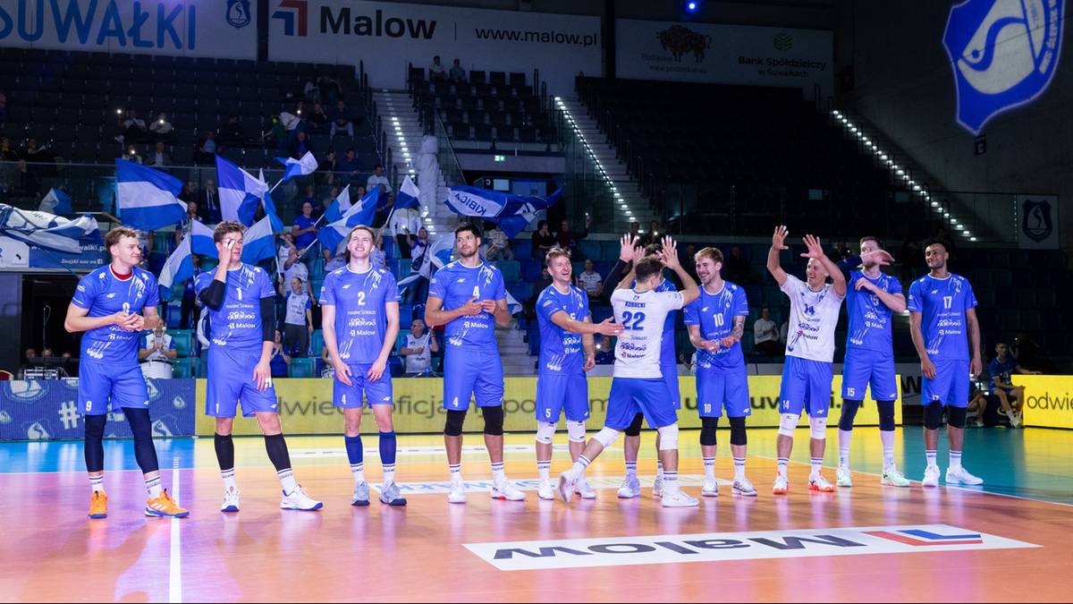 Drużyna siatkarska Ślepsk Malow Suwałki w niebieskich strojach pozuje do zdjęcia na parkiecie hali sportowej.