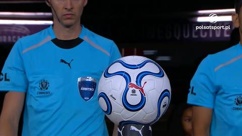 Piłka nożna marki Puma z niebieskimi wzorami na białym tle, umieszczona na cokole. W tle widoczni są dwaj mężczyźni w niebieskich koszulkach sędziowskich.