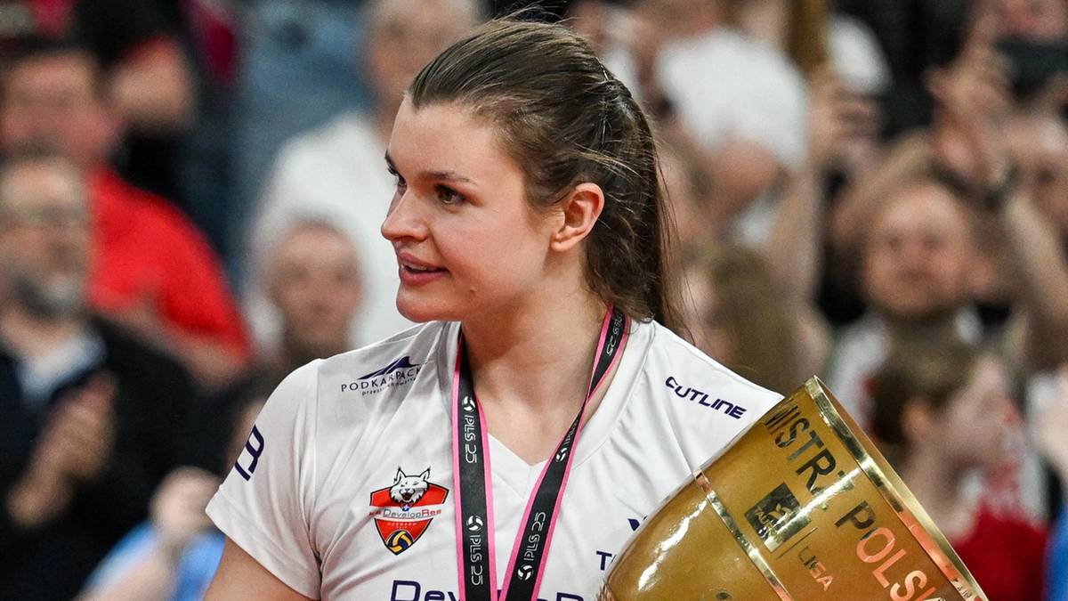 Sięgająca po trofeum młoda kobieta, siatkarka, w białej koszulce sportowej z logotypem i medalem na szyi. W tle widać rozmazane sylwetki kibiców.