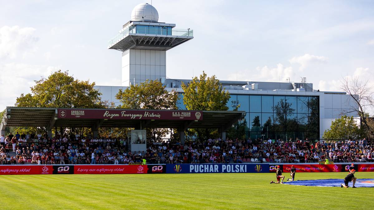 Stadion piłkarski z trybunami, budynkiem klubowym i zawodnikami na boisku.