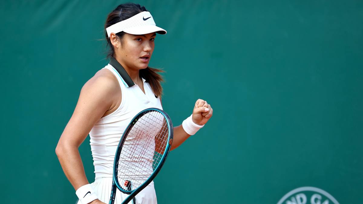 Tenisistka w białej koszulce i białej czapce z daszkiem, trzymająca rakietę tenisową w prawej ręce, z zaciśniętą lewą pięścią uniesioną w geście radości.