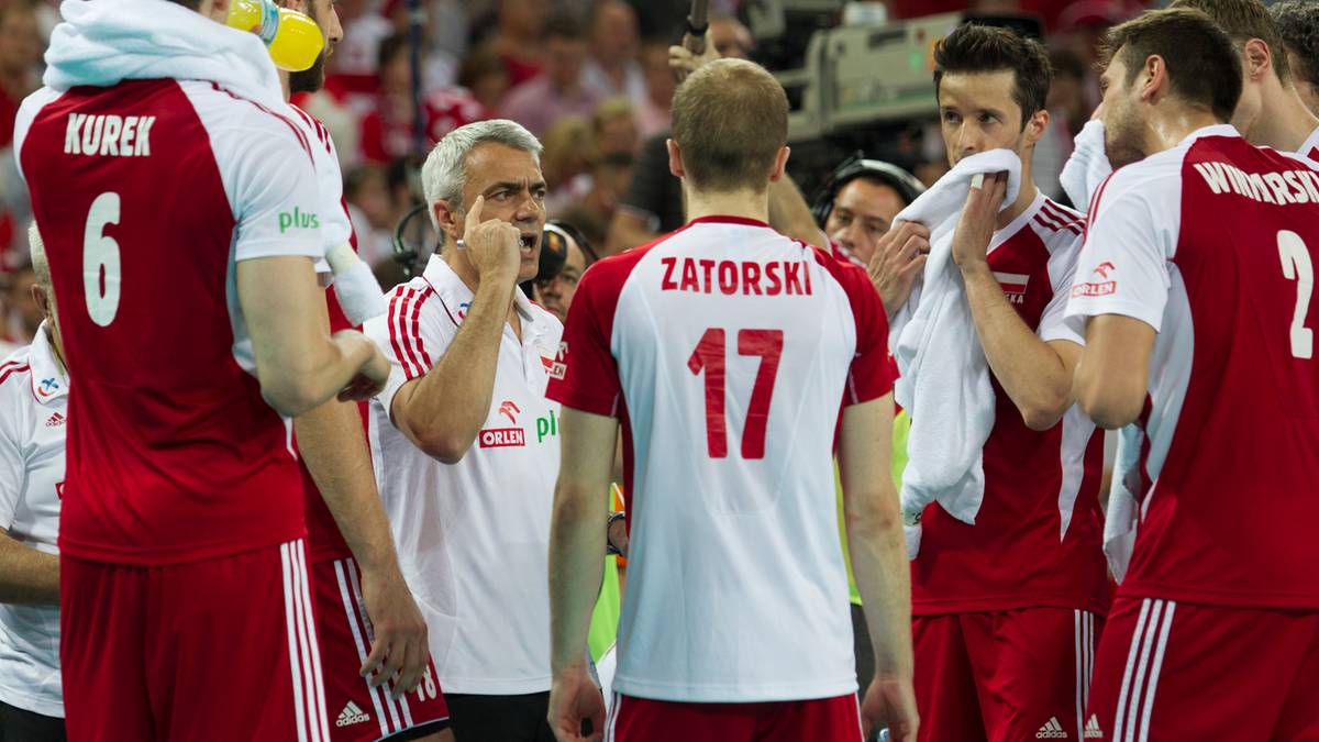 Trener w białej koszulce instruuje siatkarzy w czerwonych koszulkach z numerami na plecach.