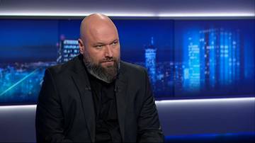 Mężczyzna z łysą głową i brodą w czarnym garniturze siedzi na tle ekranu z nocnym widokiem miasta.