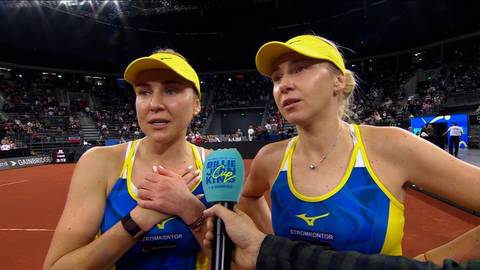 Dwie kobiety w ż&oacute;łtych czapkach z daszkiem i sportowych strojach z niebieskimi elementami, jedna z nich trzyma dłonie złożone na piersi, druga patrzy w stronę kamery, obie udzielają wywiadu na tle kortu tenisowego z widzami na trybunach.