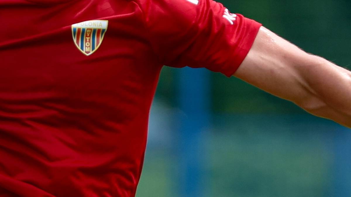 Fragment koszulki piłkarskiej w kolorze czerwonym z herbem drużyny.