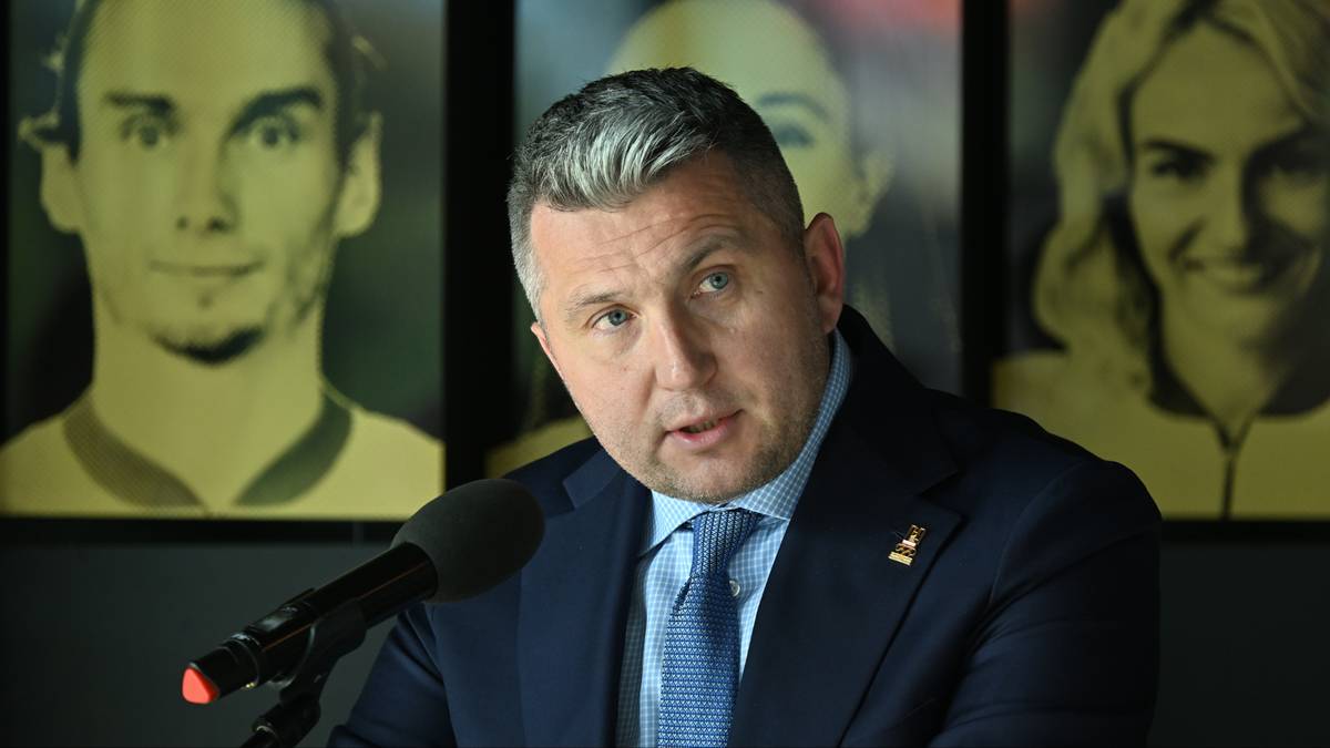 Radosław Piesiewicz w garniturze i krawacie przemawia przy mikrofonie na tle portret&oacute;w sportowc&oacute;w.