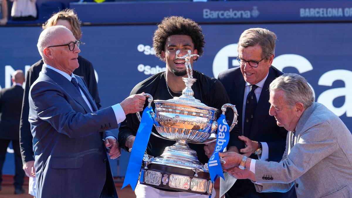 Dw&oacute;ch starszych mężczyzn wręcza puchar młodemu tenisisiście, kt&oacute;ry stoi pośrodku z uśmiechem na twarzy.