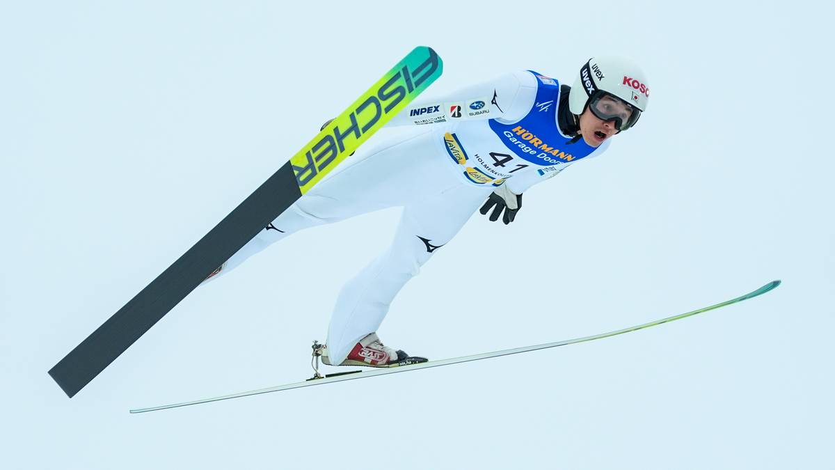Skoczek narciarski w białym kombinezonie i kasku z logo Fischer na nartach leci w powietrzu.