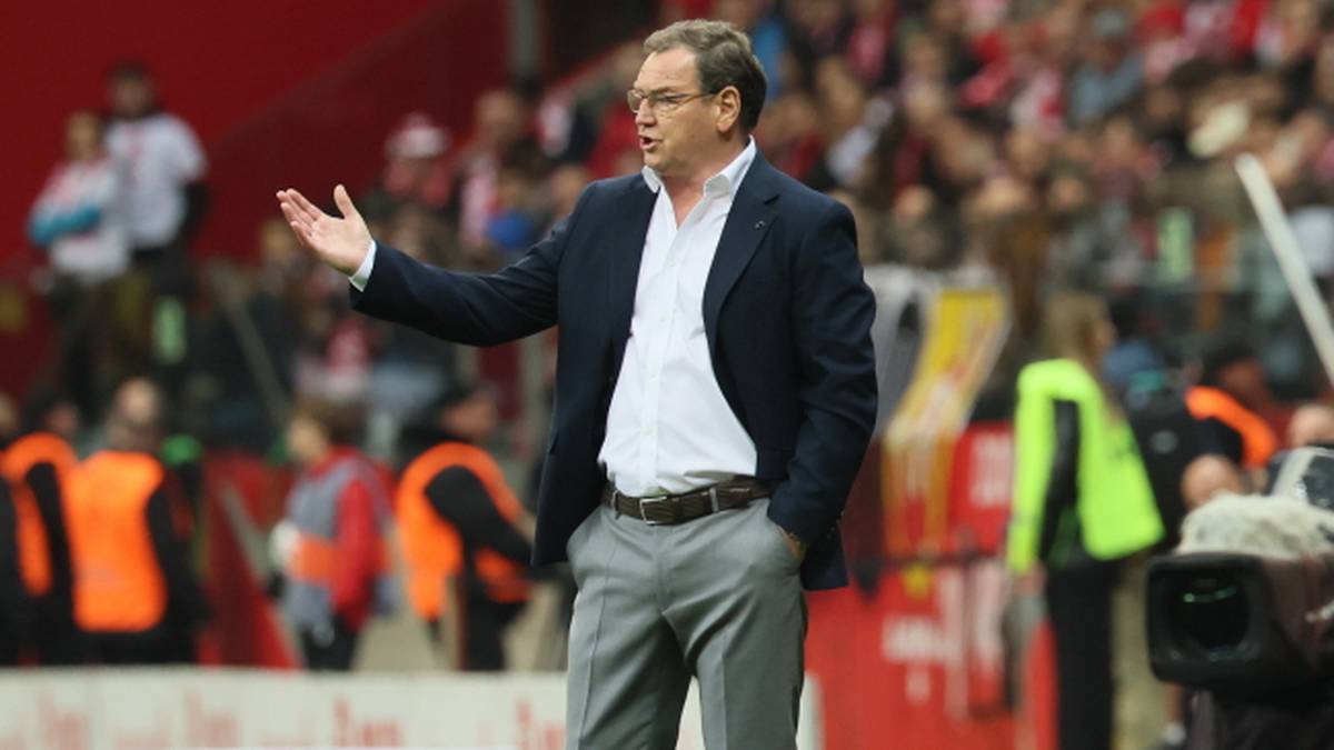 Trener piłkarski w marynarce i koszuli stoi na boisku, gestykulując prawą ręką na tle trybun stadionu.