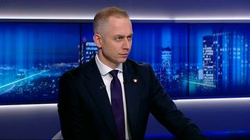 Mężczyzna w garniturze siedzi, patrząc w bok. Na jego piersi widoczna jest przypinka.