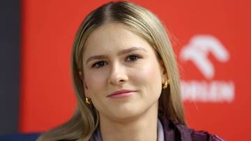 Portret kobiety z blond włosami, lekko uśmiechnięta, z widocznym logo sponsora w tle.