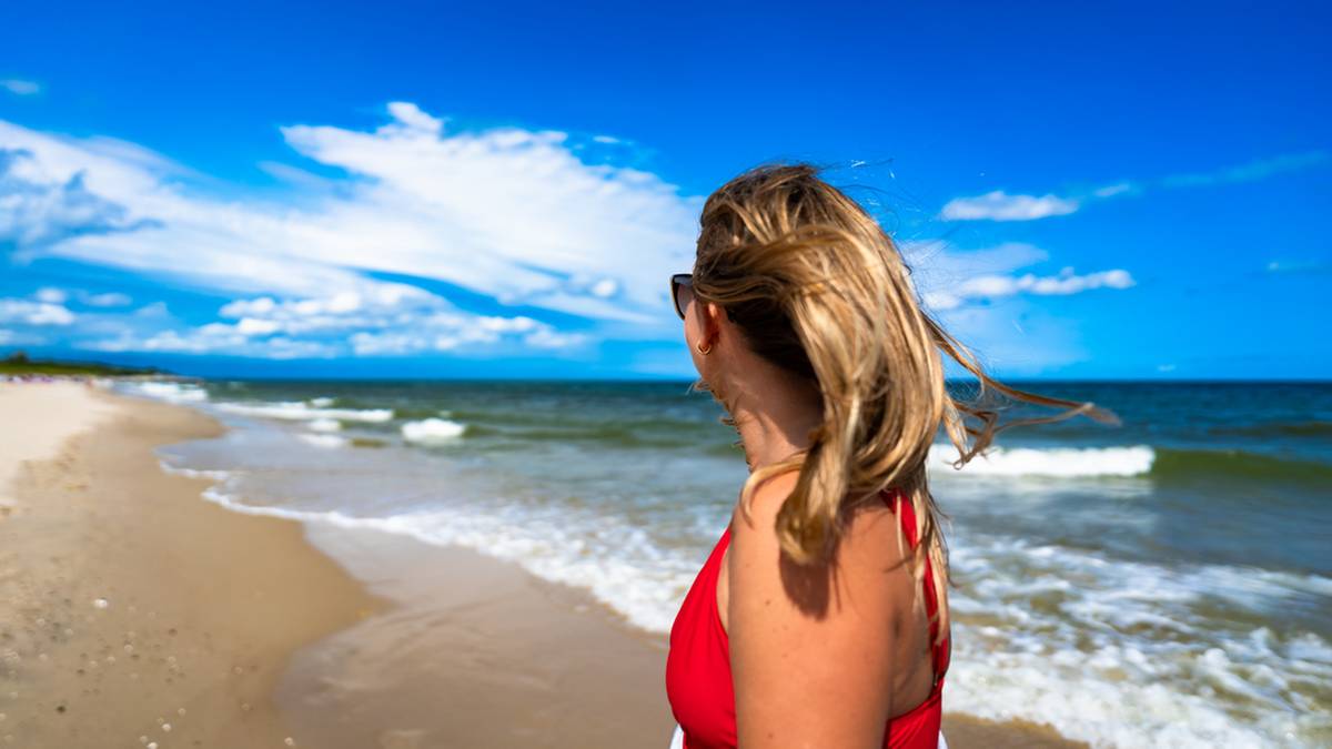 Kobieta w czerwonym stroju kąpielowym patrzy na morze, stojąc na plaży z falującymi białymi włosami.