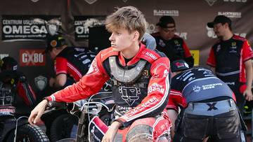 Młody żużlowiec siedzi na motocyklu, ubrany w kombinezon.