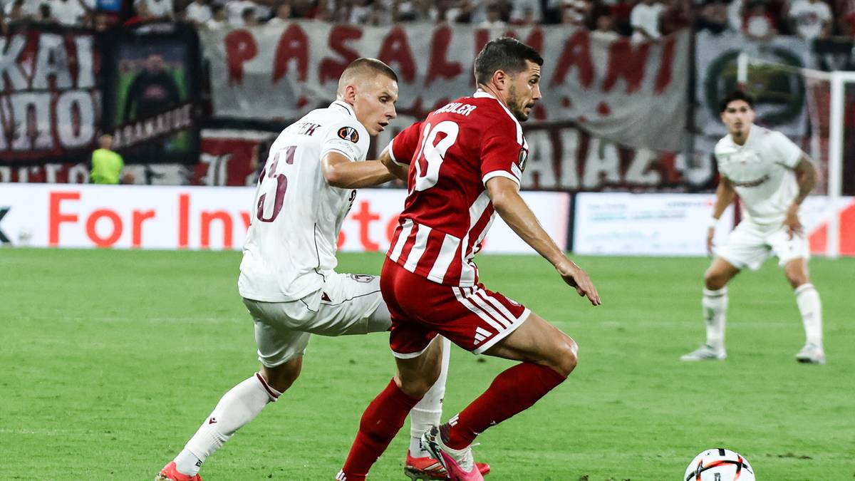 Dw&oacute;ch piłkarzy walczy o piłkę na boisku. Jeden w białym stroju, drugi w czerwonym z białymi pasami. W tle widać trybuny stadionu i innych zawodnik&oacute;w.