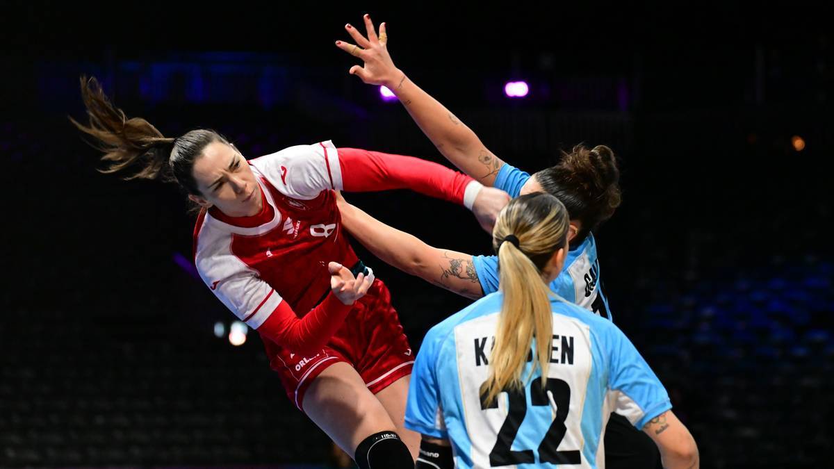 Trzy zawodniczki piłki ręcznej w akcji, jedna w czerwono-białym stroju pr&oacute;buje rzucić piłkę, dwie w niebieskich strojach pr&oacute;bują ją zatrzymać.