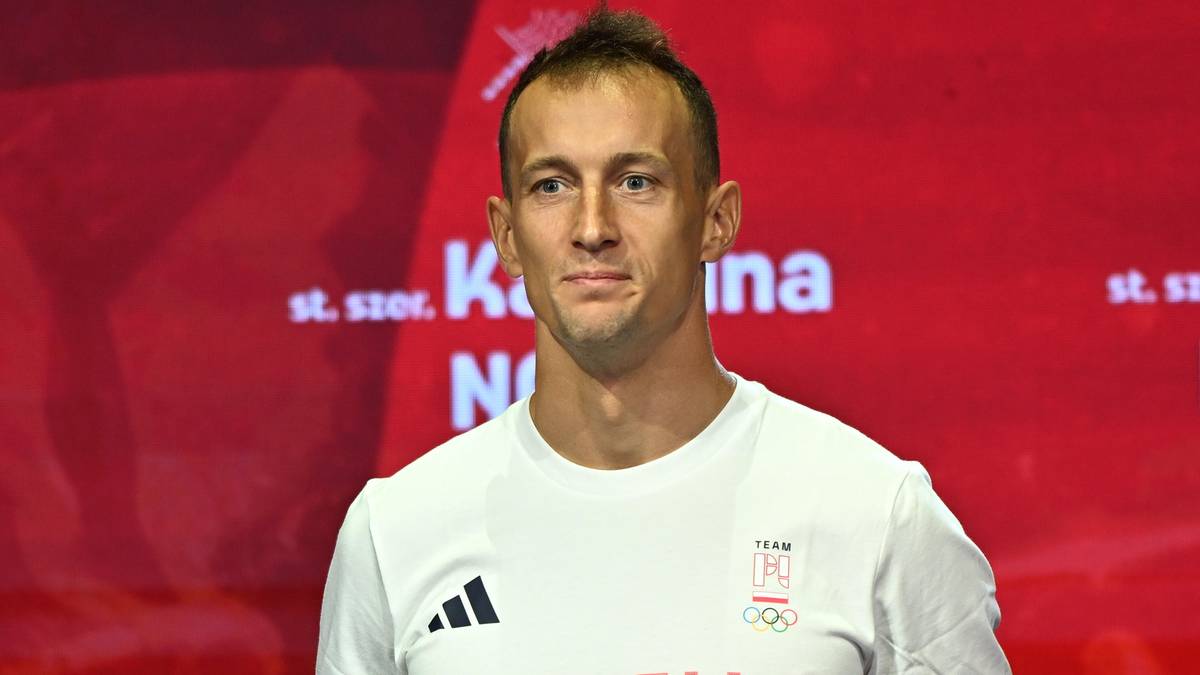 Mężczyzna w białej koszulce z napisem "TEAM POLSKA" i logiem Adidas stoi na podium, trzymając medal.