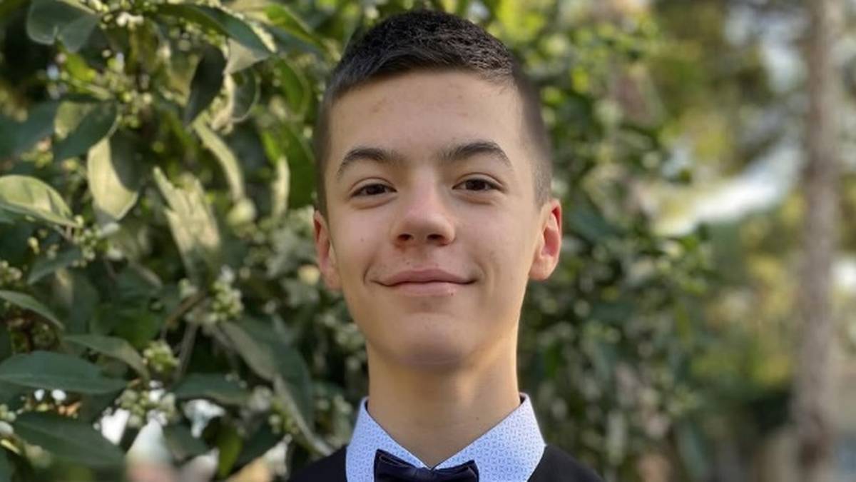 Portret młodego chłopca w czarnej muszce i białej koszuli, uśmiechającego się na tle zielonych liści.