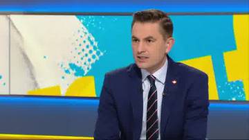 Mężczyzna w granatowym garniturze, białej koszuli i krawacie siedzi na tle studia telewizyjnego z kolorowym, geometrycznym motywem.