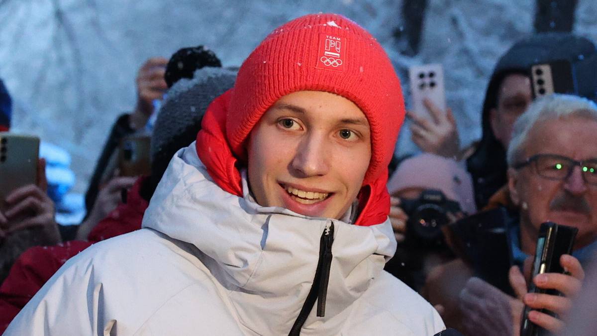 Kacper Tomasiak w białej kurtce z napisem "POLSKA" i czerwonej czapce uśmiecha się, stojąc obok młodszego chłopca, otoczeni przez tłum kibiców na zewnątrz.