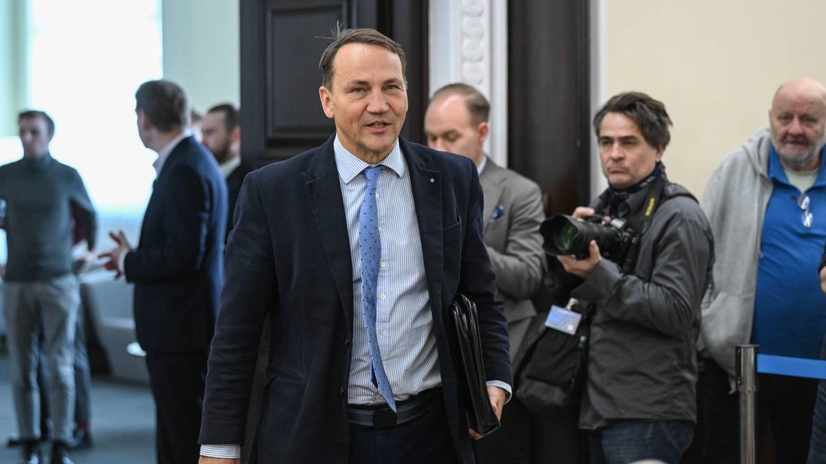 Radosław Sikorski w garniturze, otoczony przez mężczyzn i dziennikarzy.