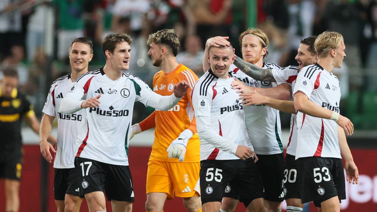 Grupa piłkarzy w białych koszulkach Legii Warszawa świętuje zwycięstwo. Jeden z piłkarzy, z numerem 17, jest w centrum uwagi, otoczony przez kolegów.