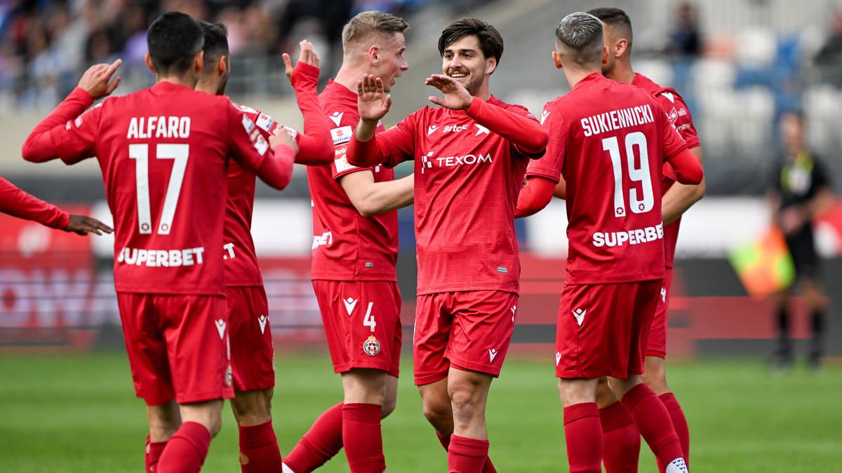 Pięciu piłkarzy w czerwonych strojach sportowych, z numerami na plecach, świętujących razem na boisku piłkarskim. Dwóch z nich ma uniesione ręce, pokazując radość.