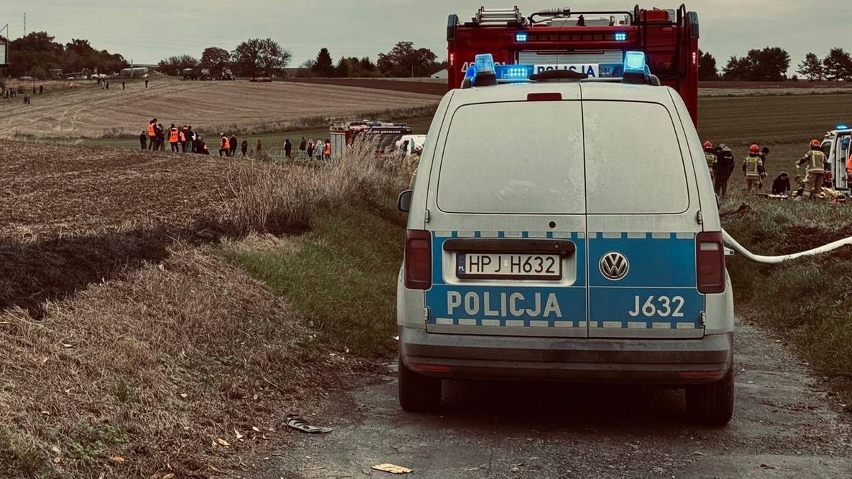 Tylna część białego radiowozu policyjnego z napisem "POLICJA" i numerem J632 na niebieskim pasie. W tle widać wóz strażacki oraz grupę osób.