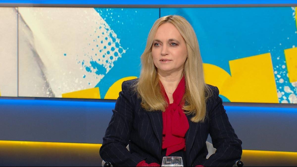 Kobieta z blond włosami, ubrana w ciemną marynarkę i czerwoną bluzkę, siedzi przed kolorowym tłem.