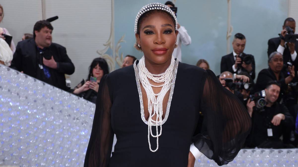 Serena Williams w czarnej sukni i białych naszyjnikach, z opaską na głowie, pozuje do zdjęcia.