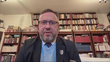 Mężczyzna w okularach i z zarostem, ubrany w marynarkę i sweter, z emblematem na piersi, mówi do kamery. Tło stanowią regały wypełnione książkami.